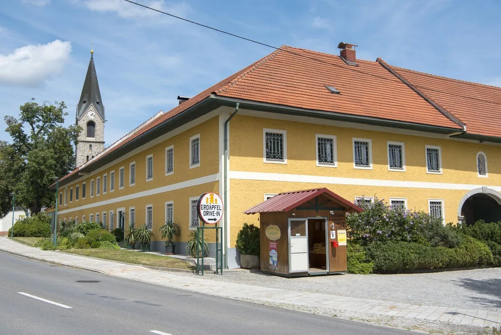 Der gelbe Vierkanthof mit rotem Ziegeldach, davor die Verkaufshütte und das Rundschild, links der Kirchturm von Pupping