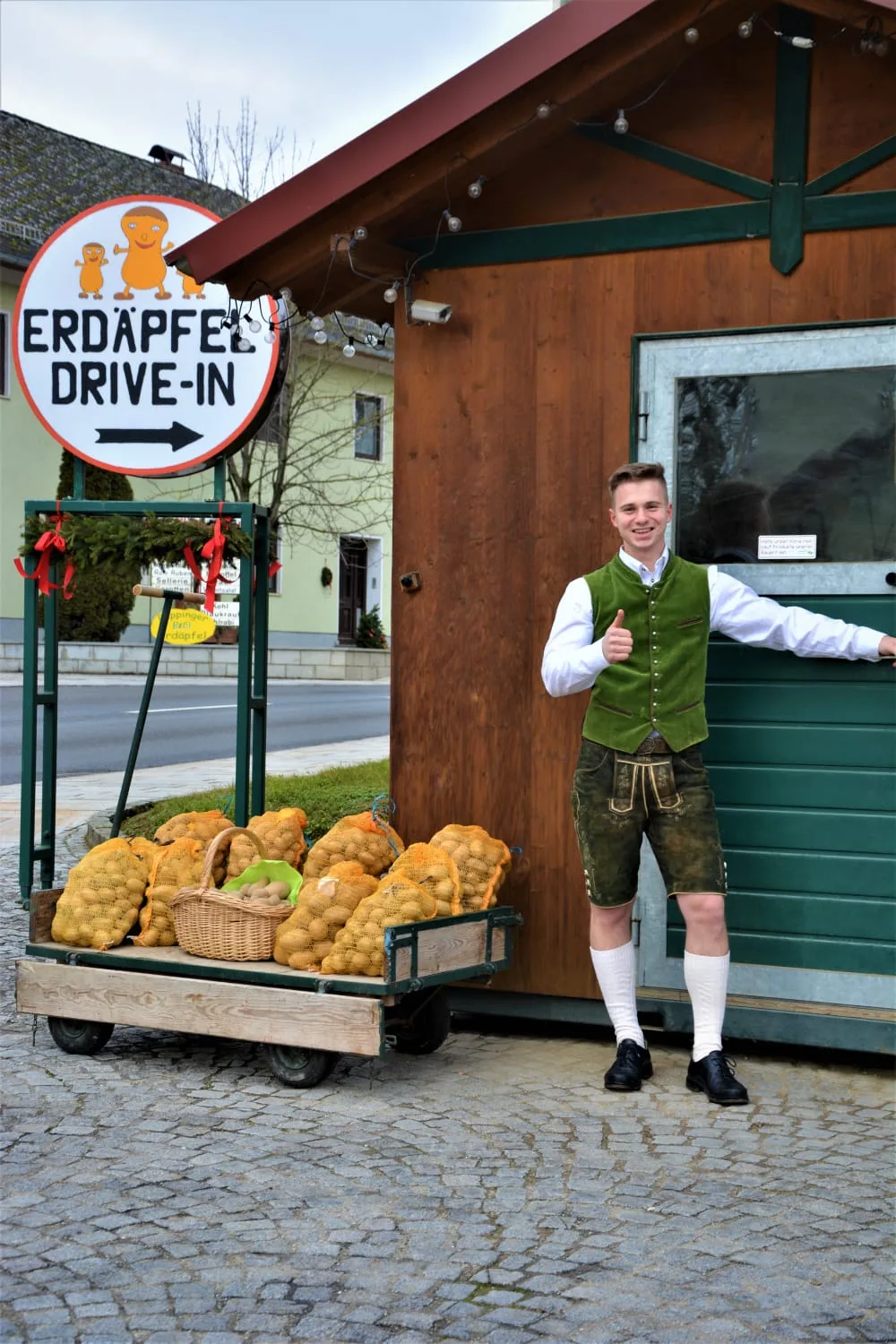 Das runde Schild Erdäpfel Drive-In über einem Leiterwagen voll Erdäpfelsäcken, daneben die Verkaufshütte