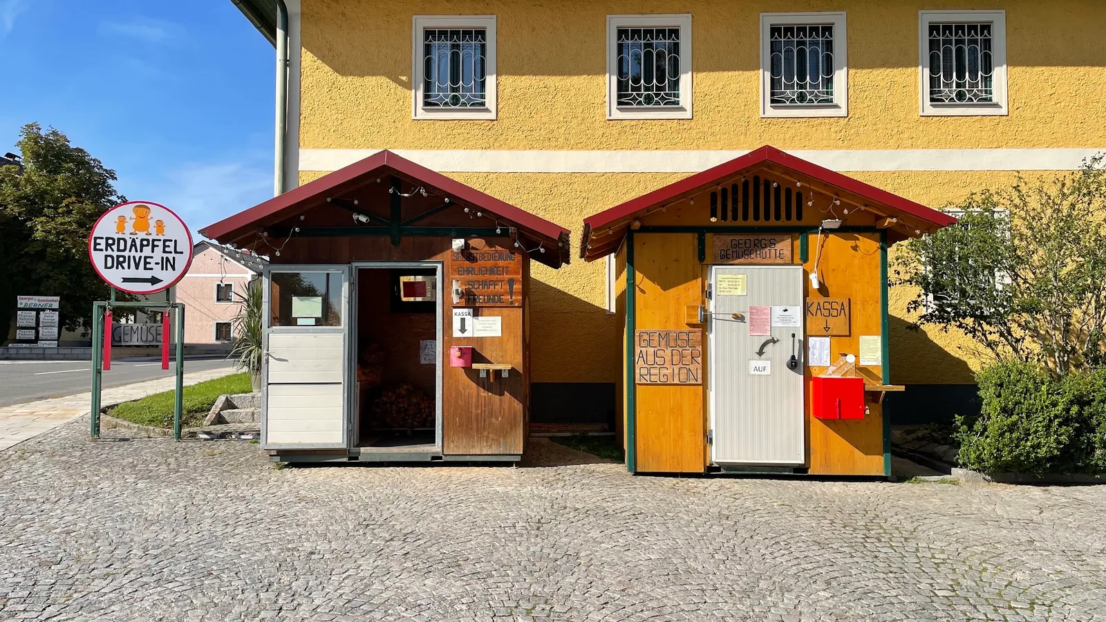 Die beiden Verkaufshütten vor der gelben Hofmauer, links das Rundschild, auf der Holzwand steht Selbstbedienung, Ehrlichkeit schafft Freunde