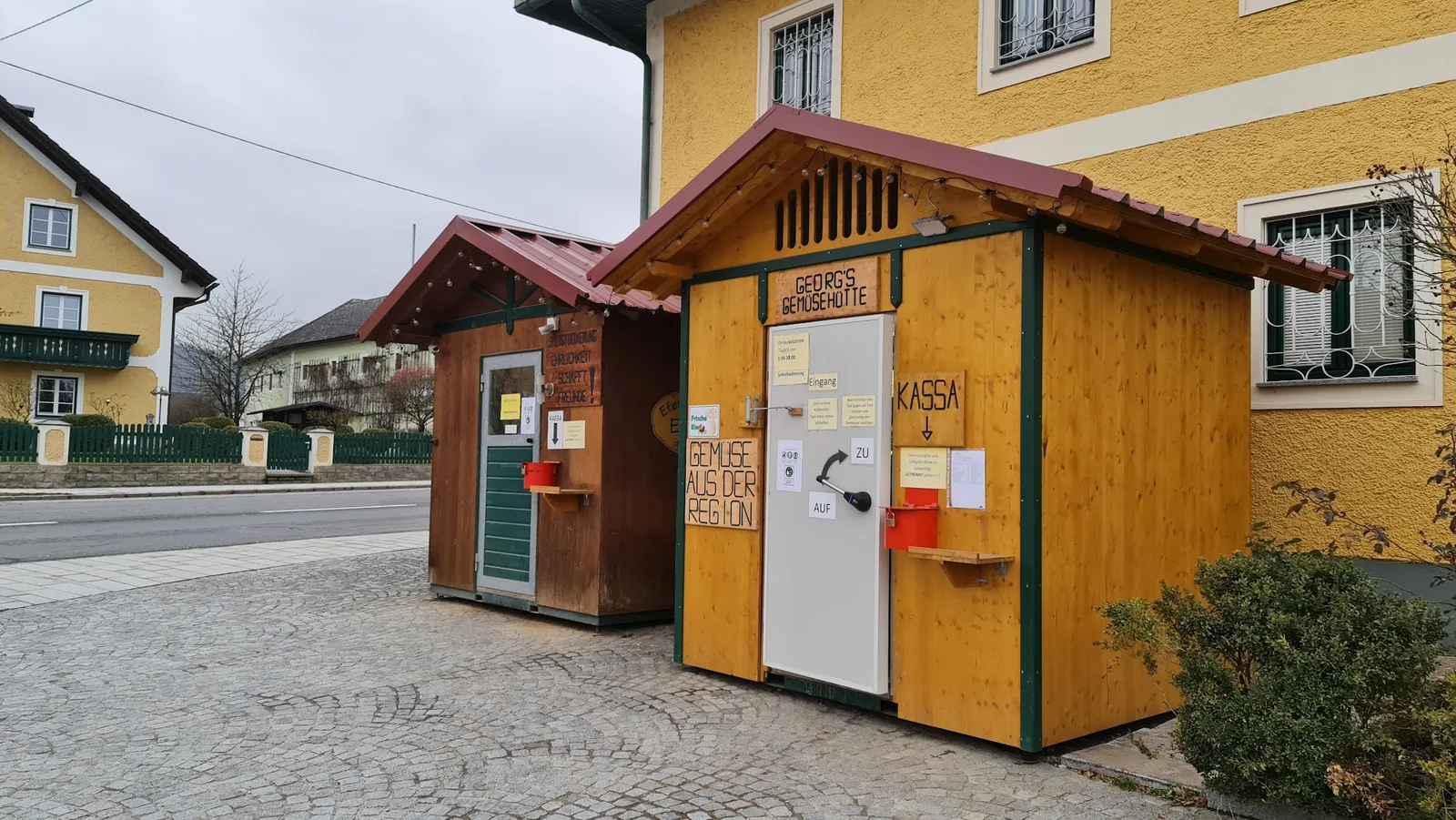 Beide Hütten an einem grauen Wintertag, auf der Türe der Gemüsehütte hängen die Öffnungszeiten und der Hinweis zur Kassa