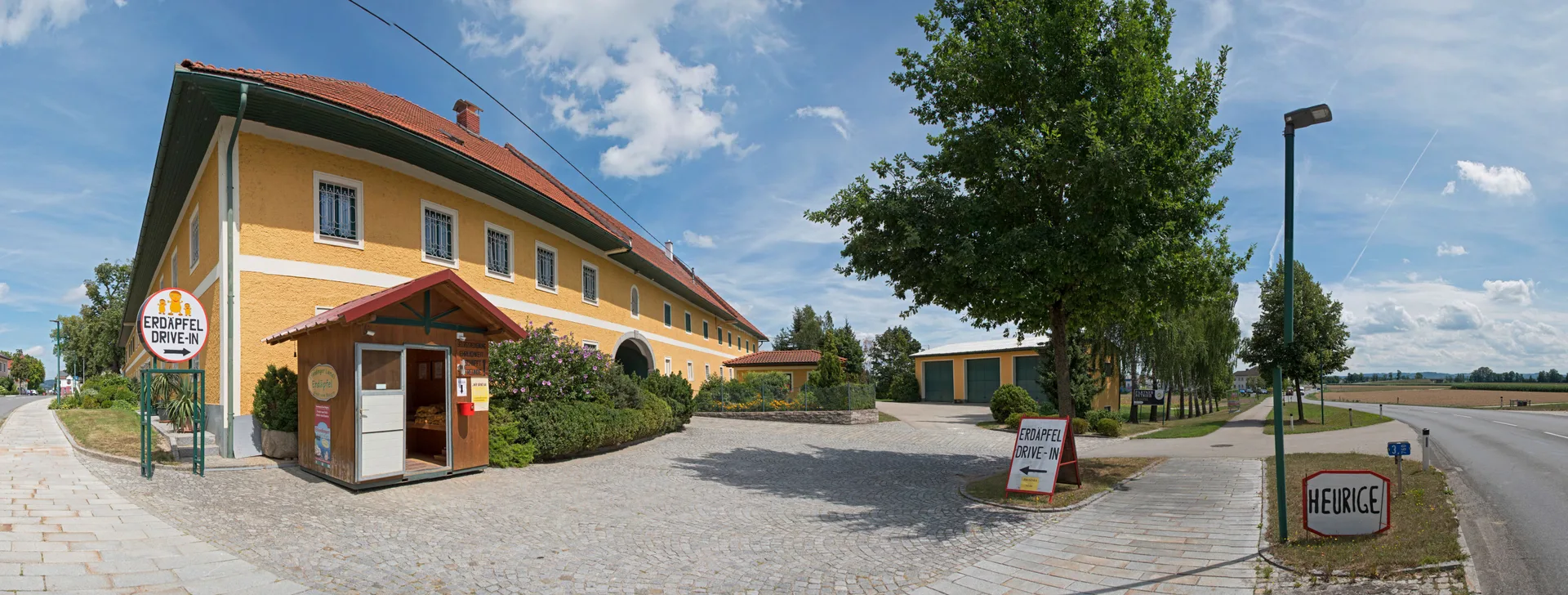 Panorama des gelben Vierkanthofs in Pupping mit der Verkaufshütte, dem Rundschild und der Straße davor