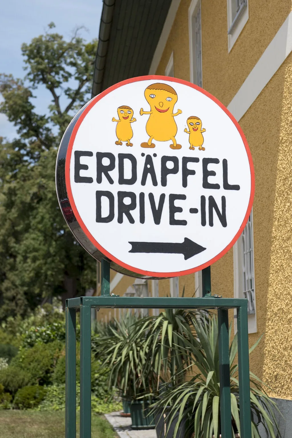 Das runde Hofschild aus der Nähe: roter Ring, drei gemalte Erdapfel-Figuren und der Schriftzug Erdäpfel Drive-In